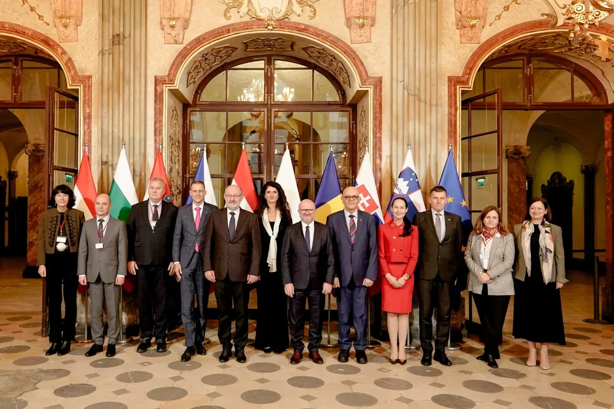 Grupa oficjalnych delegatów pozuje do pamiątkowego zdjęcia w reprezentacyjnej sali przed flagami państwowymi.