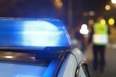 Migający niebieski sygnał świetlny na dachu radiowozu nocą, w tle rozmyta postać w kamizelce odblaskowej.