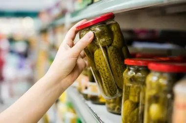 Dłoń klienta sięga po słoik ogórków kiszonych na półce w sklepie spożywczym.