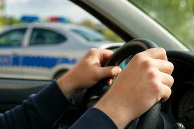 Dłonie kierowcy na kierownicy samochodu, w tle rozmyty policyjny radiowóz z włączonymi sygnałami świetlnymi.