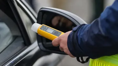Ręka policjanta w niebieskim mundurze trzymająca żółty alkomat przy otwartym oknie samochodu.